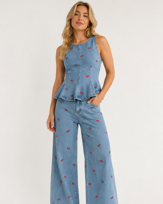 A woman modeling a light blue denim two-piece set featuring a sleeveless peplum top and matching wide-leg pants, both decorated with small red embroidered cherry motifs, set against a plain white studio background.